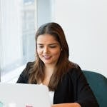 Smiling woman in black blazer working on laptop with open notebook on desk in a modern office setting.